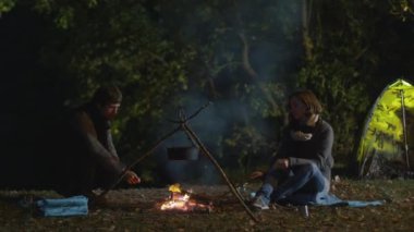 Bir erkek ve bir kadın bir kamp ateşi yanında geceleri oturup ve başka bir erkek odun ateşe atar iken bir sopa yemek hazırlamak.
