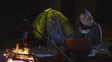 Genç esmer kadın geceleri bir kamp ateşi yanında bir laptop kullanıyor.