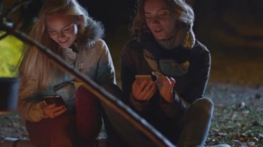 İki mutlu kız gece bir kamp ateşi yanında oturan ve onların akıllı telefonlar kullanmak.
