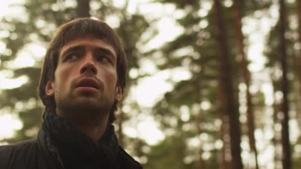 Длинноволосый мужчина в темной осенней одежде и шарфе стоит в лесу и смотрит вокруг . — стоковое видео