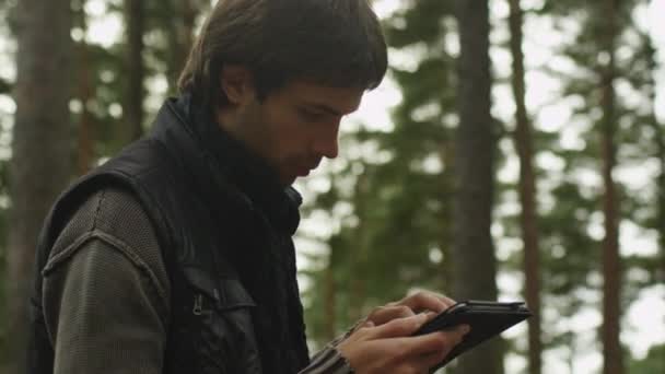 Hombre de pelo largo en ropa oscura de otoño y bufanda está de pie en un bosque y utilizando una tableta . — Vídeos de Stock