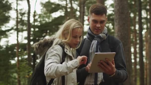 Short haired man and blond woman in autumn clothes are standing in a forest and using tablet. — Stock Video