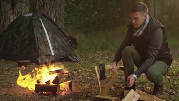Homem de cabelos curtos em roupas de outono é cortar madeira com um machado e jogá-lo no fogo . — Vídeo de Stock