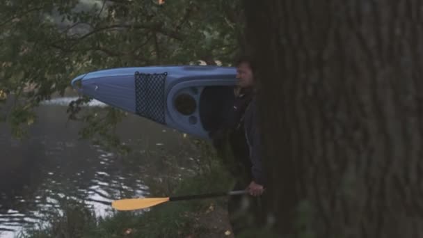 El hombre está llevando un kayak azul a la orilla del río y lo pone en el agua . — Vídeos de Stock