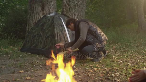 El hombre está montando una tienda en un bosque mientras dos chicas están sentadas al lado del fuego . — Vídeos de Stock