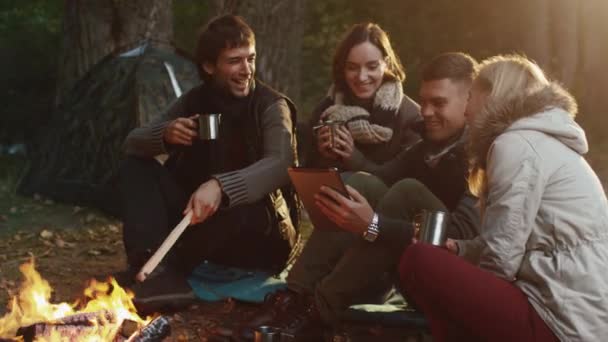 Skupina mužů a žen, sedět u táborového ohně s nápoji a v lese při použití tabletu. — Stock video