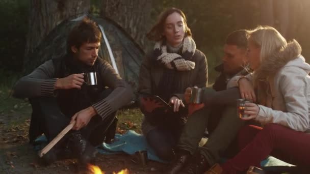 Skupina lidí sedí v lese u táboráku s teplé nápoje z termosky a při použití tabletu. — Stock video