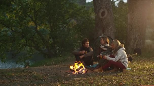 Grupo de hombres y mujeres se sientan en un bosque junto a una fogata con bebidas calientes del termo y hablan . — Vídeos de Stock