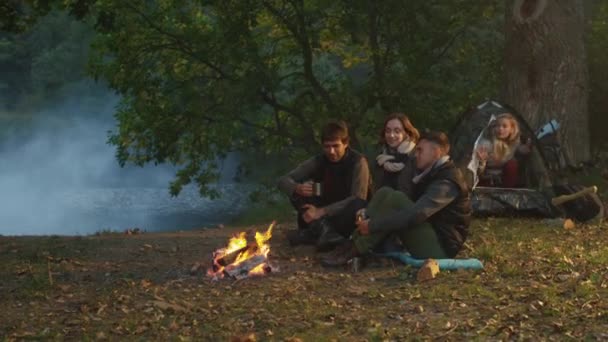 Group of friends sit in a forest near a lake next to a campfire with warm drinks while another girl steps out from a tent. — Stock Video