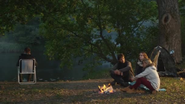 Group of friends sit in a forest next to a campfire with warm drinks and talk while another man sits in a chair near a lake. — Stock Video
