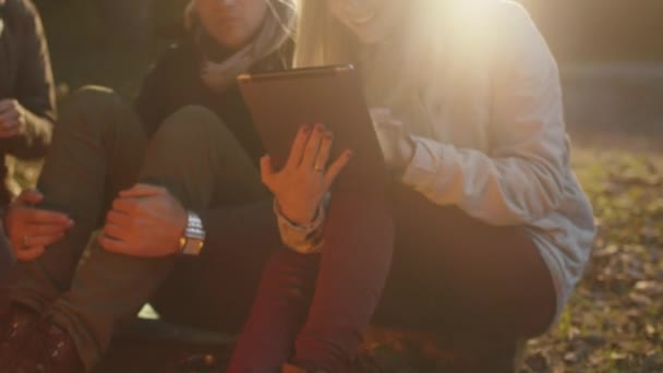 Happy young woman is using a tablet next to a campfire and showing the screen to friends. — Stock Video