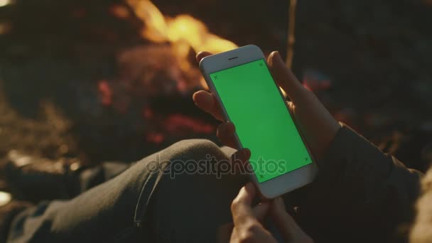 Το κορίτσι κρατά ένα smartphone σε λειτουργία πορτρέτου με πράσινη οθόνη mock-up δίπλα σε ένα campfire το βράδυ. — Αρχείο Βίντεο
