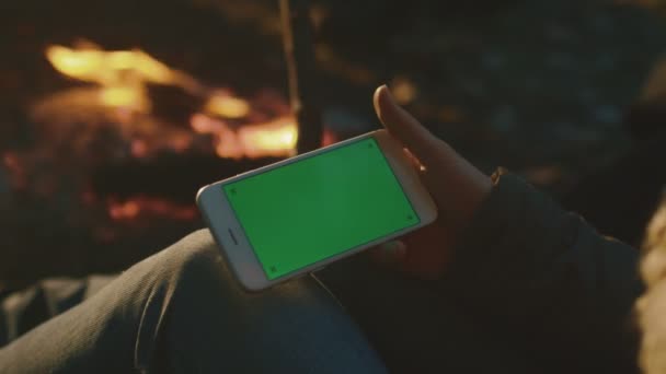 Chica está sosteniendo un teléfono inteligente en modo paisaje con pantalla verde maqueta al lado de una fogata por la noche . — Vídeo de stock