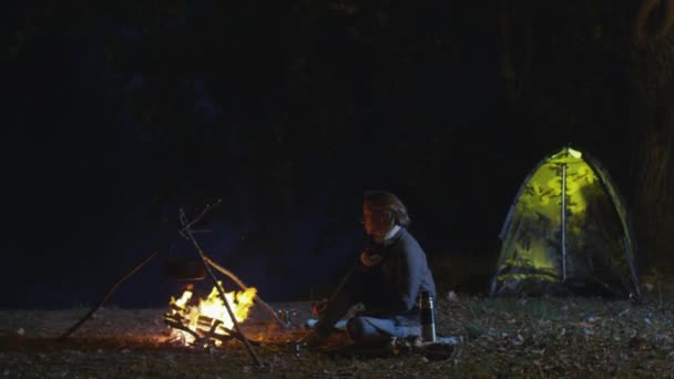 女孩正坐在一堆篝火旁边晚上带个帐篷在背景中. — 图库视频影像