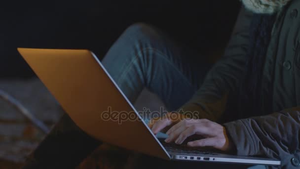 Jonge brunette vrouw is het gebruik van een laptop naast een kampvuur bij nacht in close-up. — Stockvideo