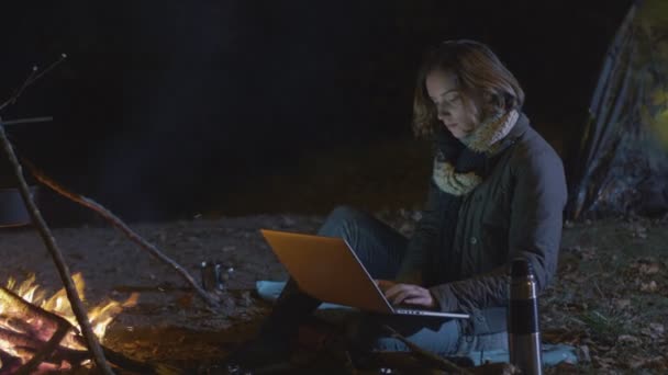 Junge brünette Frau benutzt einen Laptop neben einem Lagerfeuer in der Nacht. — Stockvideo