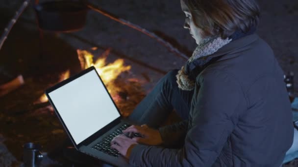 Bruna ragazza sta usando un computer portatile con un modello di schermo accanto a un falò di notte . — Video Stock