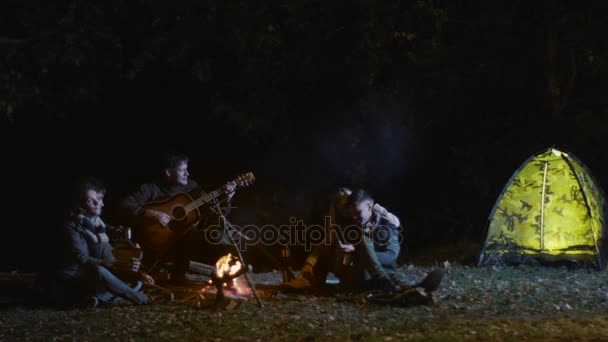 El hombre está tocando en una guitarra al lado de la fogata por la noche mientras un grupo de amigos están escuchando . — Vídeos de Stock