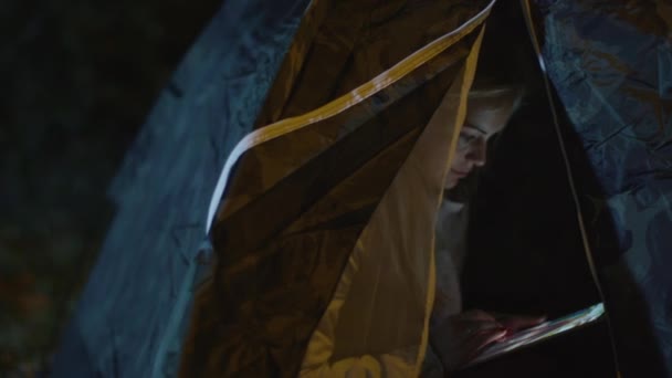 Jeune fille blonde utilise une tablette dans une tente la nuit . — Video