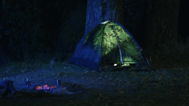 A luz é apagada em uma tenda ao lado de uma fogueira em chamas à noite em uma floresta . — Vídeo de Stock