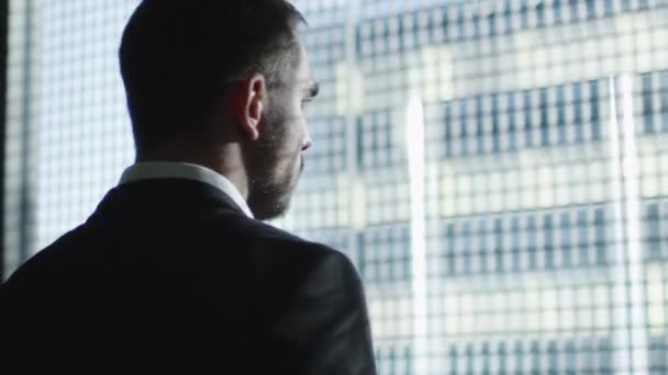 Uomo d'affari in giacca e cravatta guarda fuori da una grande finestra in una città in una giornata di sole . — Video Stock