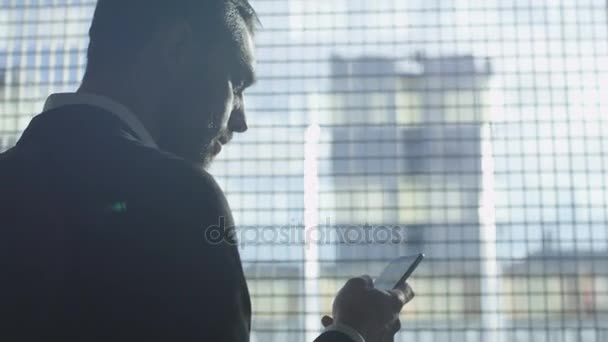 Empresario en un traje está utilizando un teléfono inteligente al lado de una gran ventana en un garaje . — Vídeos de Stock