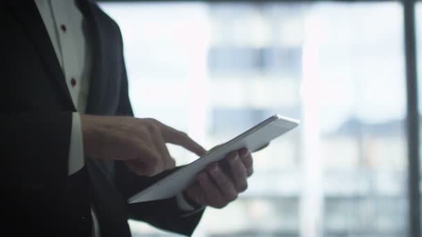 Close-up van een zakenman in een pak met behulp van een tablet naast een groot raam. — Stockvideo