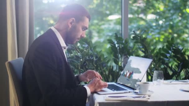 Un homme d'affaires en costume travaille sur un ordinateur portable et boit du café dans un restaurant . — Video