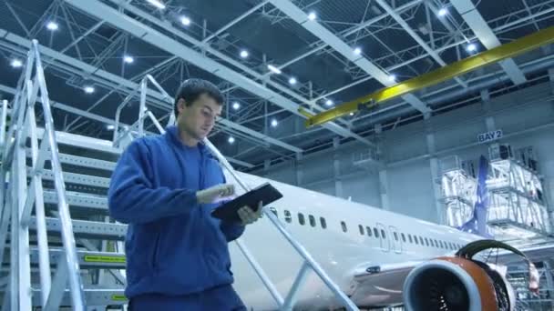 Vliegtuig onderhoud monteur gaat de trap af terwijl met behulp van tablet en groeten van zijn collega een de bodem in een hangar. — Stockvideo