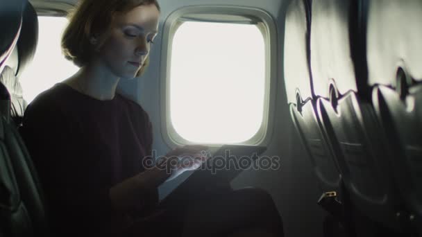 Ung kvinna använder en surfplatta inuti ett flygplan bredvid ett fönster. — Stockvideo
