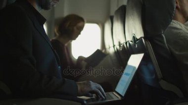 Genç adam bir uçakta bir dizüstü bilgisayar üzerinde çalışıyor ve bir kadın bir pencere yanında arka planda okuyor.
