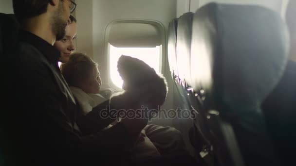 Familia joven con un niño sentado dentro de un avión al lado de una ventana y jugar con un oso de peluche . — Vídeo de stock