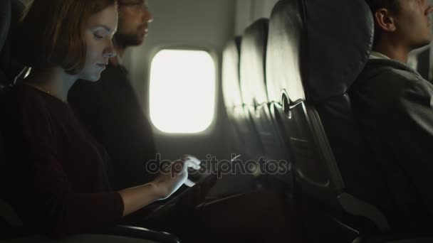 Νεαρή γυναίκα χρησιμοποιώντας ένα tablet σε ένα αεροπλάνο και έναν άνδρα είναι να ακούτε μουσική στο παρασκήνιο δίπλα σε ένα παράθυρο. — Αρχείο Βίντεο