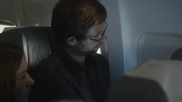 Молодой человек открывает окно в самолёте и смотрит на улицу, в то время как рядом с ним сидит женщина. . — стоковое видео