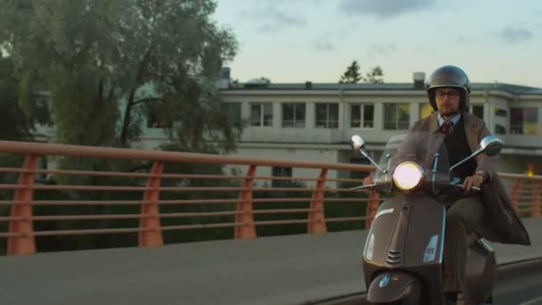 Jonge man in een elegante beige trenchcoat rijdt een retro scooter over een brug bij zonsondergang. — Stockvideo