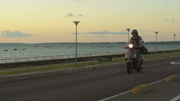 Jovem em um elegante casaco bege cavalga uma scooter retro em uma estrada à beira-mar ao pôr-do-sol . — Vídeo de Stock