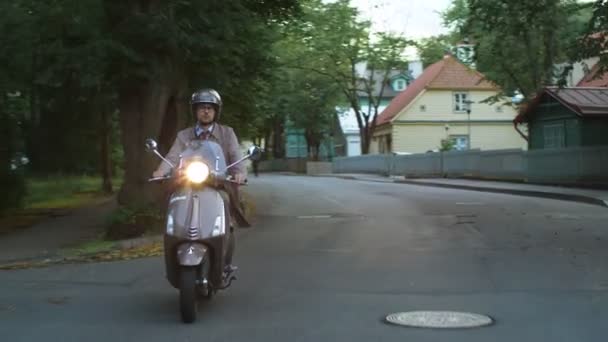 Joven en una elegante gabardina beige monta un scooter retro en una zona residencial rural . — Vídeos de Stock