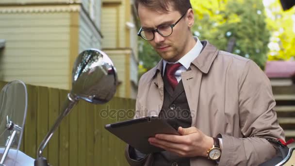 Young man in an elegant beige trench coat sits on a scooter and uses a tablet. — Stock Video