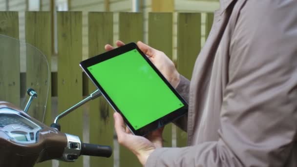 Jovem em um elegante casaco bege trincheira senta-se em uma scooter e mantém um tablet com tela verde . — Vídeo de Stock