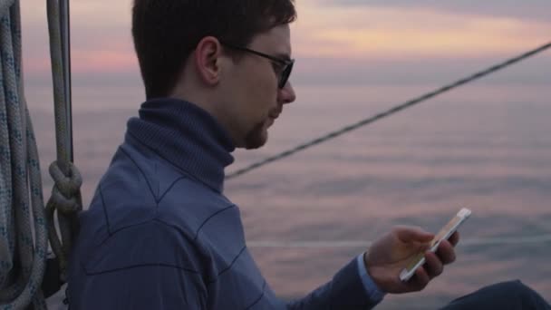Ο άνθρωπος ψάχνει και χρήση του τηλεφώνου σε ένα ιστιοπλοϊκό σκάφος στη θάλασσα στο ηλιοβασίλεμα. — Αρχείο Βίντεο