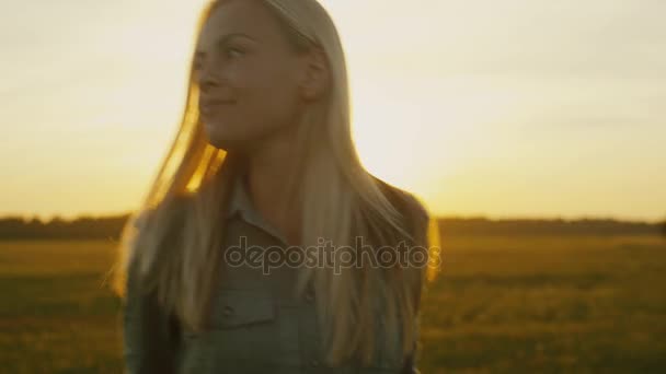 Blond meisje glimlachend loopt op veld bij zonsondergang — Stockvideo