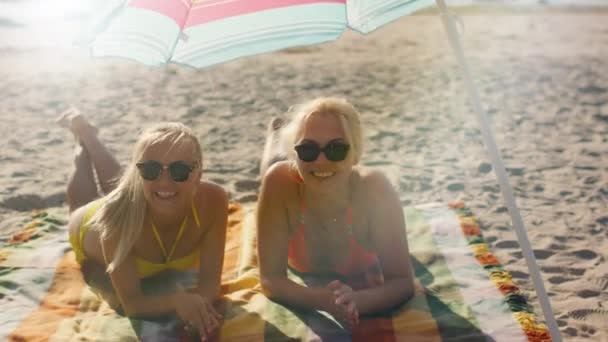 Dos chicas sonrientes están acostadas bajo paraguas en la playa y conversan . — Vídeos de Stock