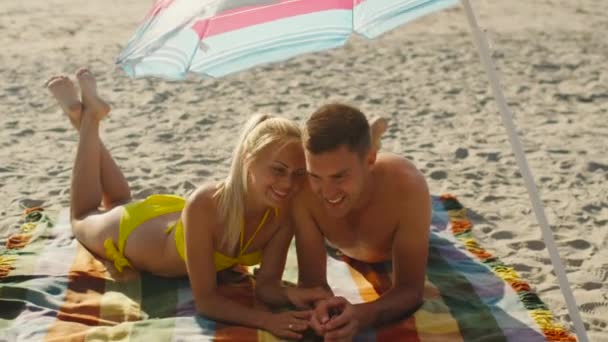 Joven pareja sonriente acostada bajo paraguas en la playa y hablando . — Vídeo de stock