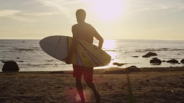 Jovem com prancha de surf está correndo para a água em luz do pôr do sol — Vídeo de Stock