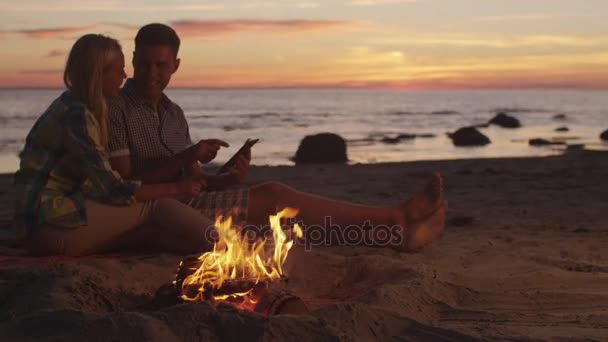 Pareja está sentada cerca de Campfire y usando Tablet PC por la noche — Vídeos de Stock