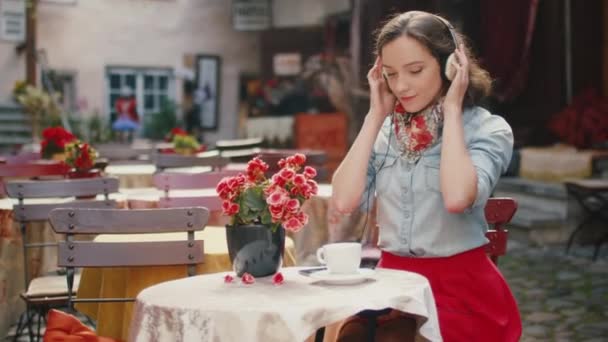 Chica escuchando música en auriculares en la cafetería al aire libre — Vídeos de Stock