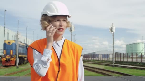 Mulher de chapéu duro e colete de segurança andando e conversando no telefone na estação ferroviária . — Vídeo de Stock