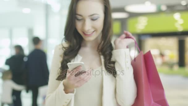 Chica morena joven con hermosa sonrisa perfecta está utilizando el teléfono móvil en el centro comercial. Disparo de mano . — Vídeos de Stock