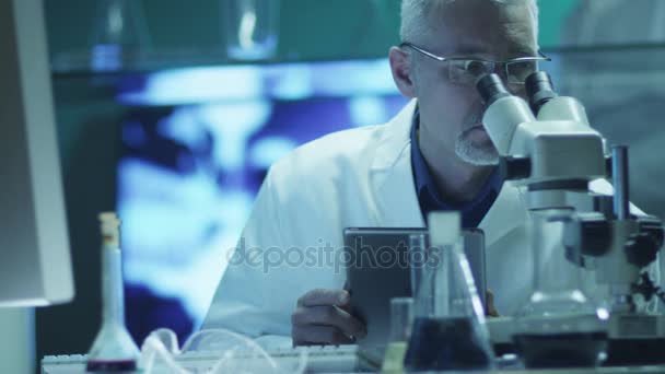 Naukowiec jest patrząc przez Mikroskop i za pomocą tabletu w laboratorium — Wideo stockowe