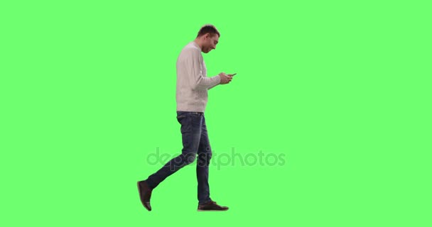 Hombre caucásico casual está caminando y utilizando el teléfono inteligente en una pantalla verde simulada en el fondo . — Vídeo de stock